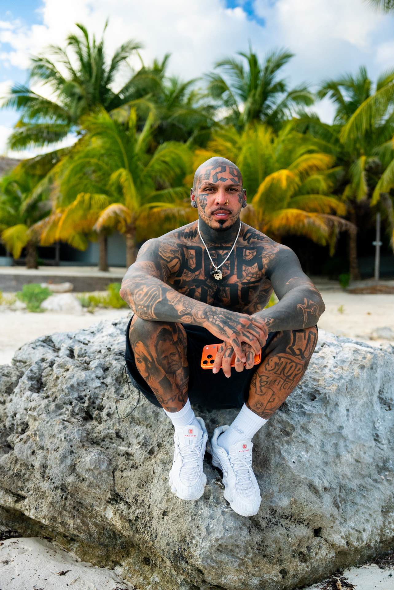 Tattooed artist seated on a coastal rock beneath palm trees