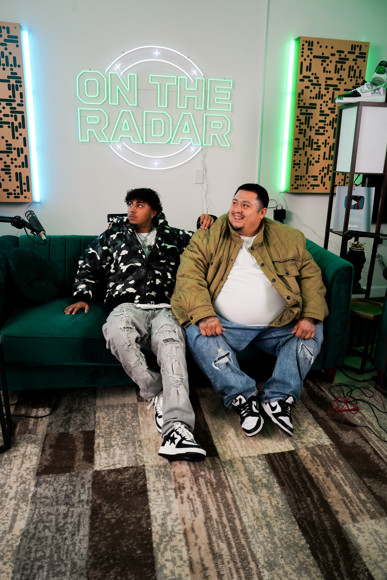 Two men seated on a green couch under an On The Radar neon sign