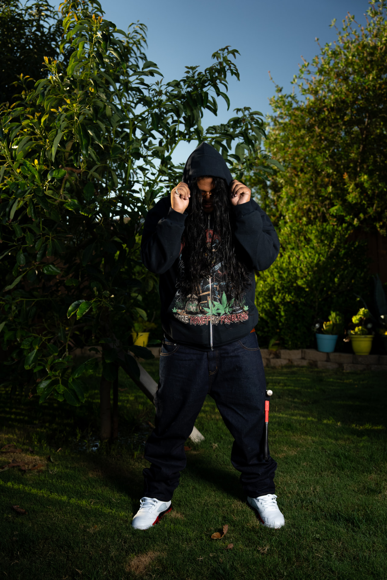 Artist pulling up a black hoodie in a sunlit backyard
