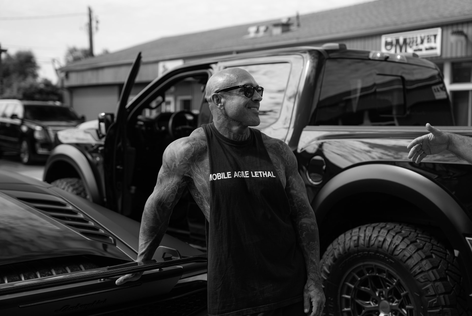 Black-and-white photo of a tattooed man beside a lifted truck and sports car